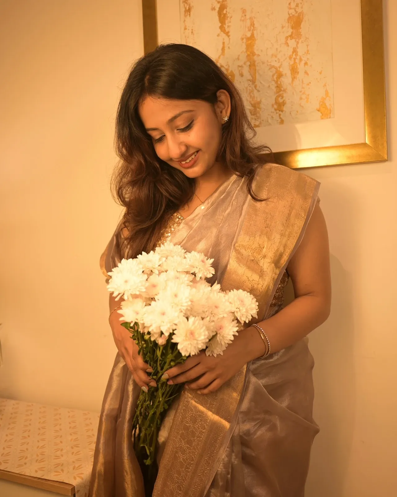 Woman in a grey gold tissue saree smiling down at a bunch of white chrysanthemums